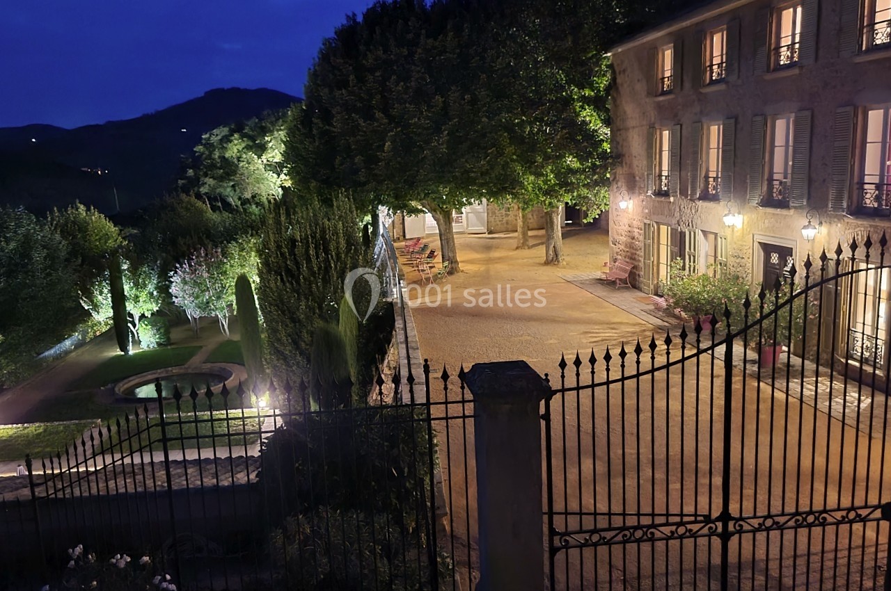 Cour d'une maison éclairée la nuit, avec jardin arboré, bassin et collines en arrière-plan.