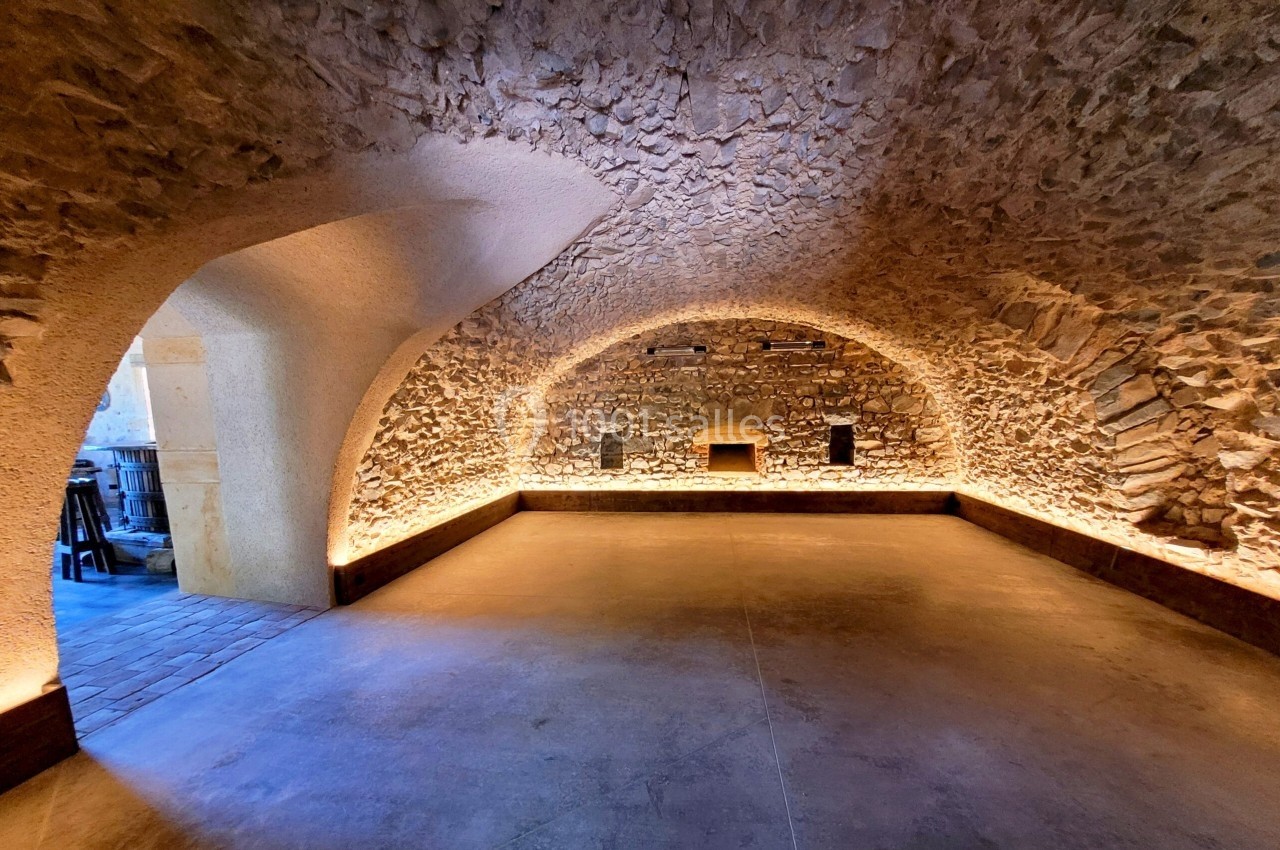 Salle voûtée en pierre avec éclairage indirect au sol, sol en béton lisse et ouverture vers un espace adjacent.
