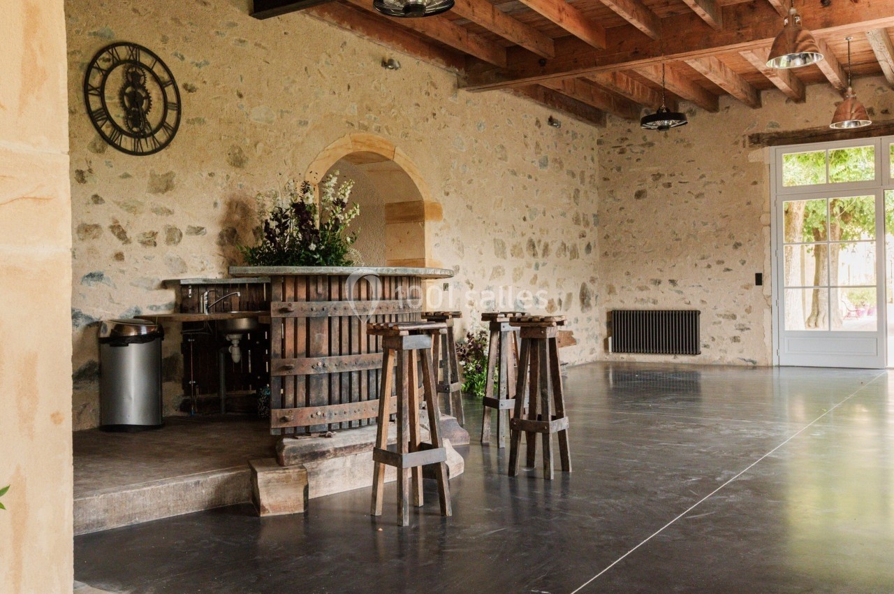 Salle avec murs en pierre, bar en bois, tabourets hauts, plafond avec poutres apparentes et porte vitrée donnant sur l…