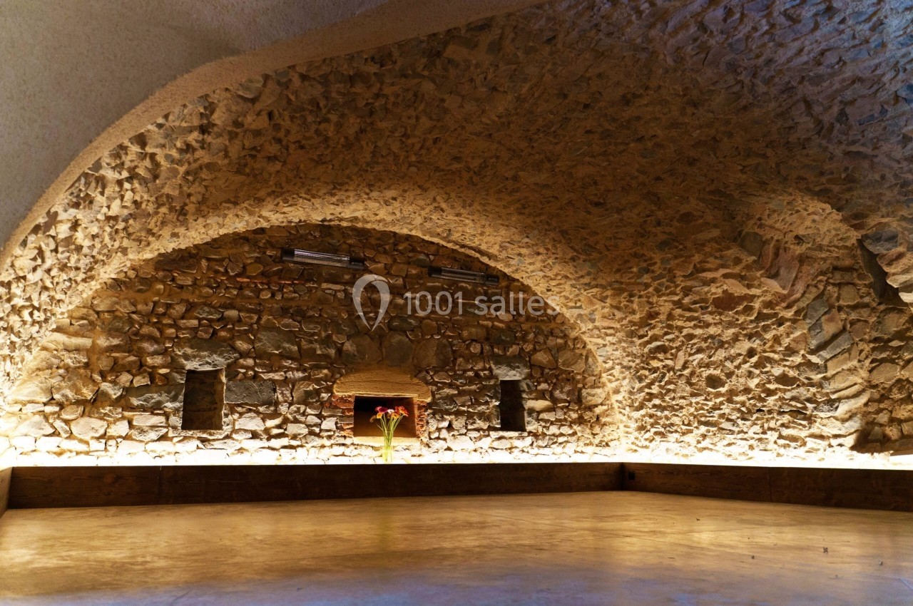 Salle voûtée en pierre avec un sol en béton poli, éclairée par des lumières chaudes, et un vase de fleurs au centre.