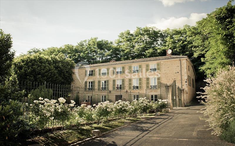 Grande maison en pierre entourée d'un jardin fleuri, avec une clôture en fer forgé et des arbres en arrière-plan.