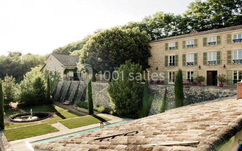 Vue d'une maison en pierre entourée de jardins aménagés avec une fontaine centrale et des arbres.