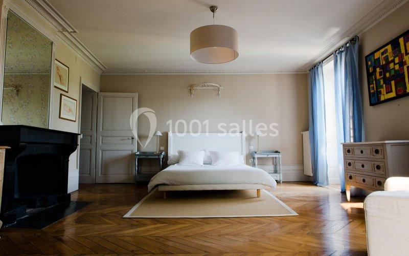 Chambre lumineuse avec lit double, parquet en bois, cheminée noire, rideaux bleus et mobilier clair.