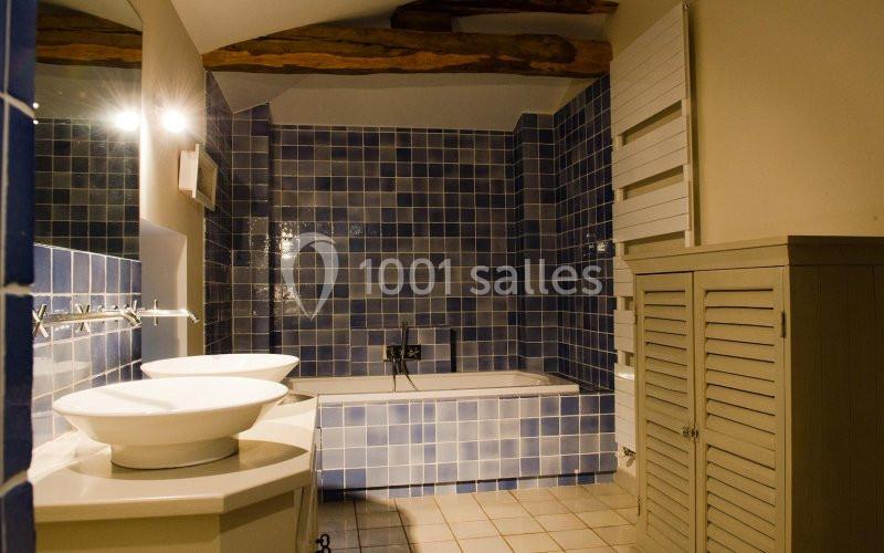Salle de bain avec carrelage bleu, baignoire encastrée, double vasque blanche et poutres apparentes au plafond.