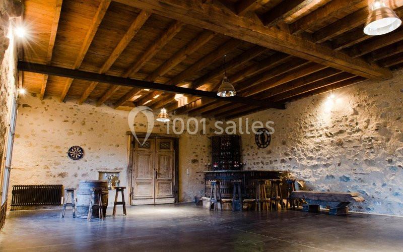 Salle rustique avec murs en pierre, plafond en bois, bar en bois, tabourets, tonneaux et éclairage suspendu.