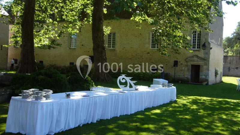 Table dressée avec nappes blanches dans un jardin verdoyant, devant un bâtiment en pierre avec des arbres ombragés.