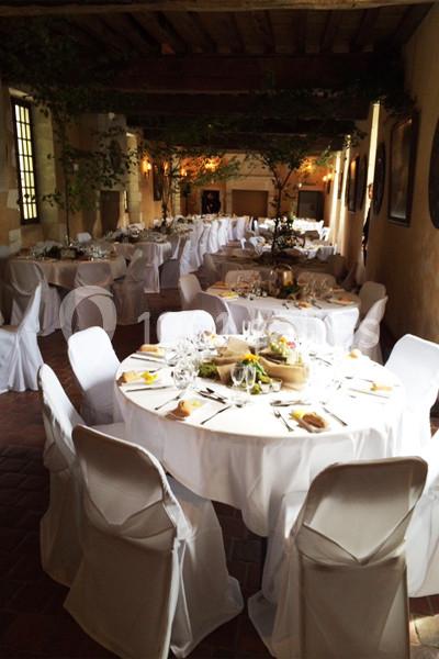 Salle de réception décorée avec des tables rondes dressées, nappes blanches et chaises houssées, éclairage chaleureux.