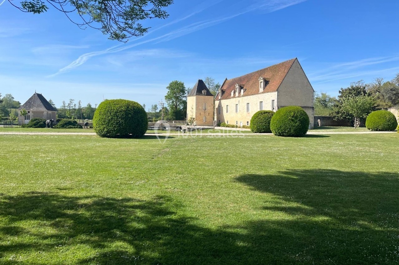 Grand jardin verdoyant avec pelouse, arbustes taillés et bâtiments en pierre sous un ciel bleu dégagé.