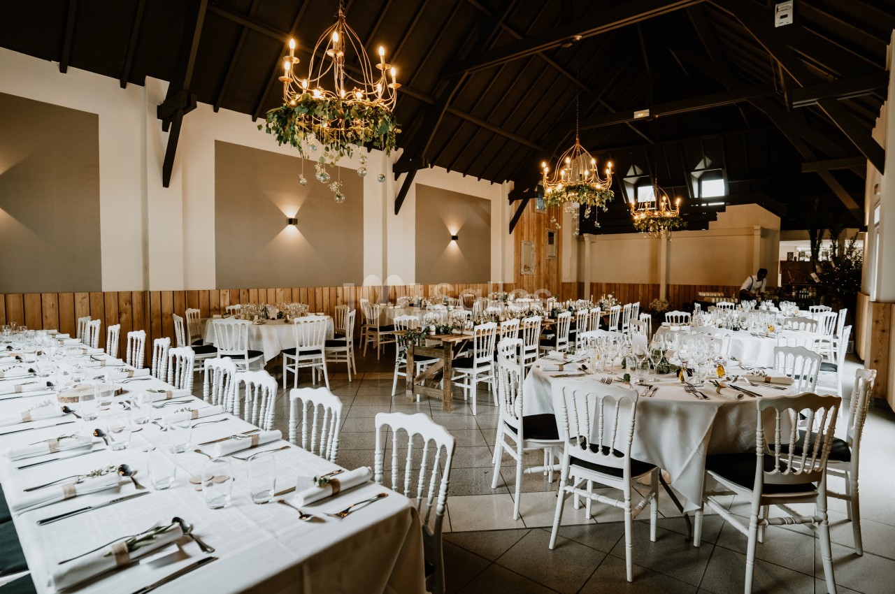 Salle de réception élégante avec tables dressées, chaises blanches et lustres suspendus dans un décor chaleureux.