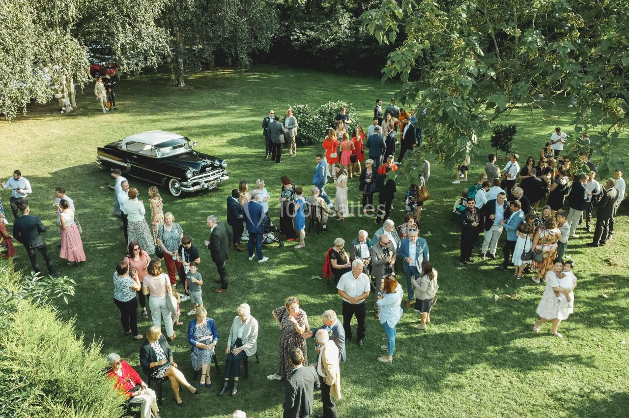 Groupe de personnes rassemblées dans un jardin verdoyant, avec une voiture ancienne noire en arrière-plan.