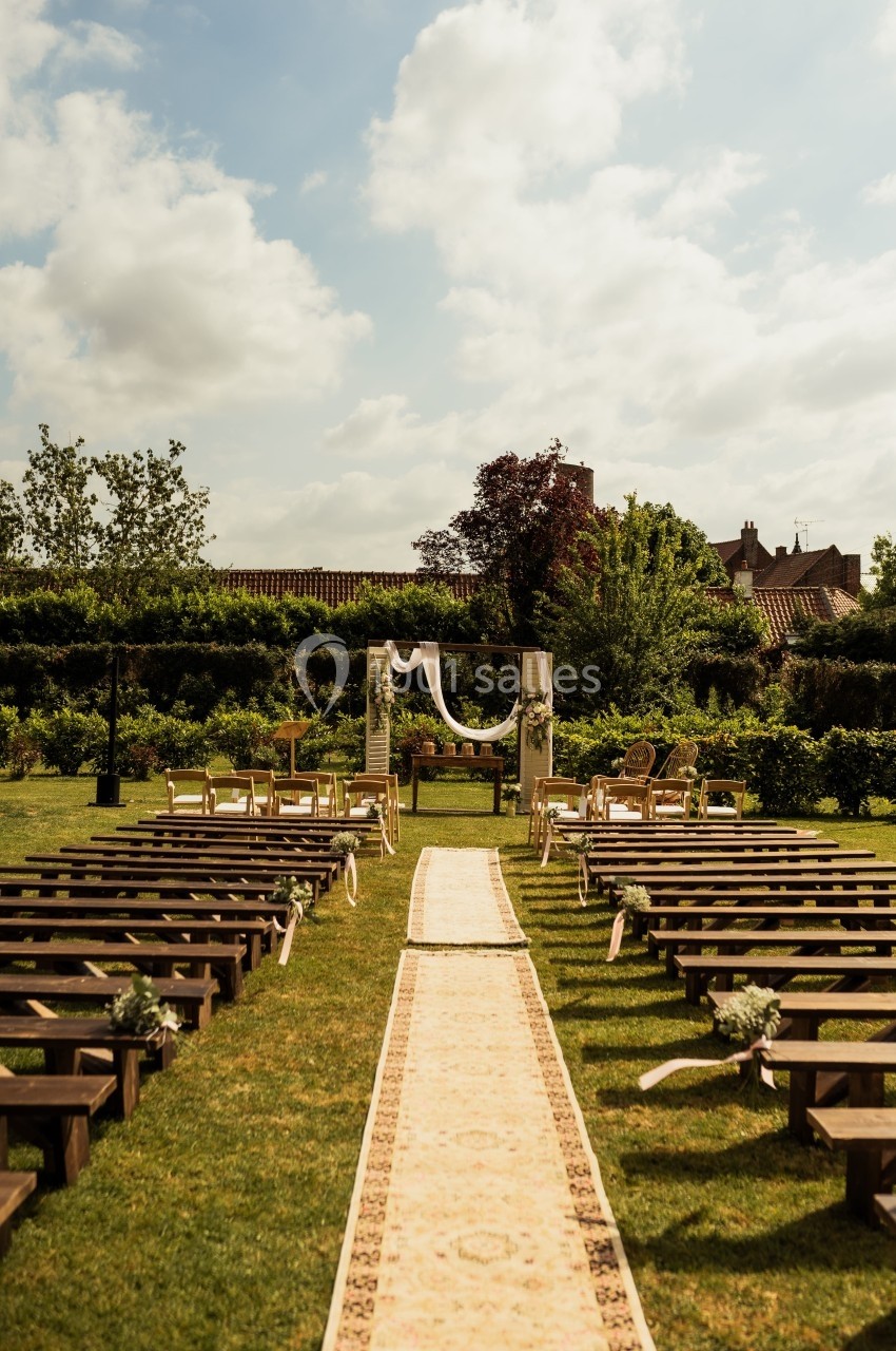 Allée centrale décorée menant à une arche de mariage entourée de bancs en bois dans un jardin ensoleillé.