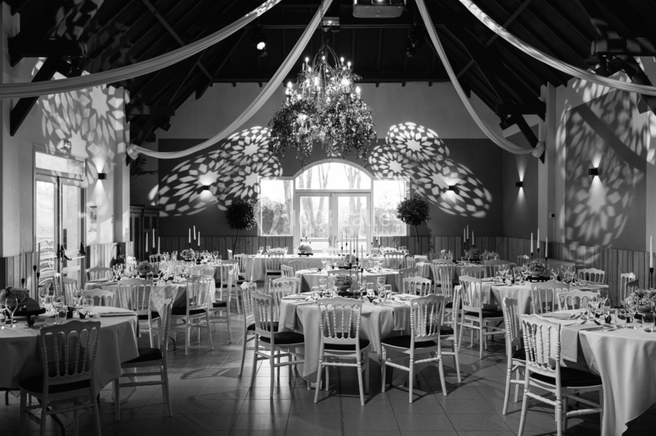 Salle de réception décorée avec des tables rondes dressées, des chaises blanches et un éclairage tamisé sous un lustre.