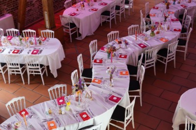 Salle de réception décorée avec des tables rondes dressées, des fleurs colorées et un mur de briques apparentes.
