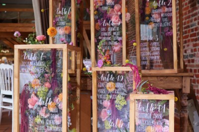 Salle de réception décorée avec des tables rondes dressées, des fleurs colorées et un mur de briques apparentes.