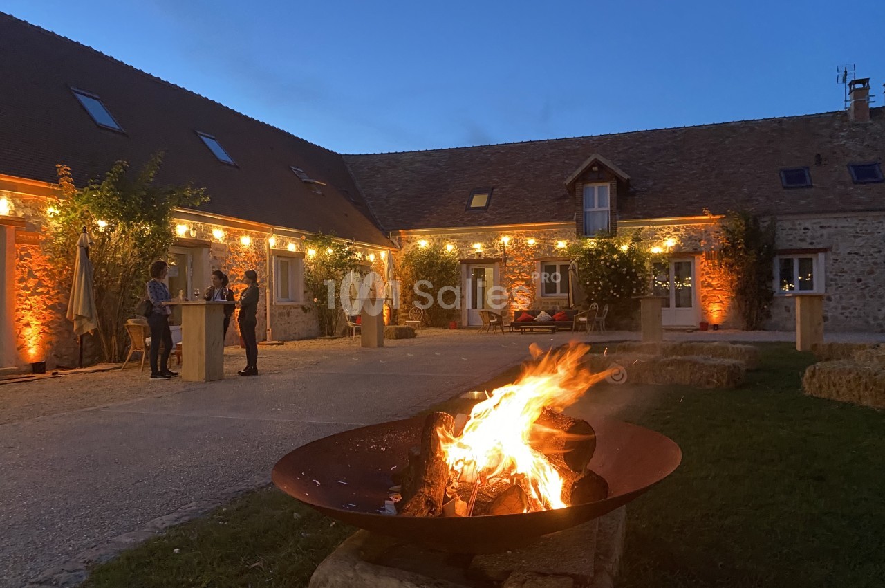 Brasero allumé dans une cour pavée entourée de bâtiments en pierre éclairés par des guirlandes lumineuses.