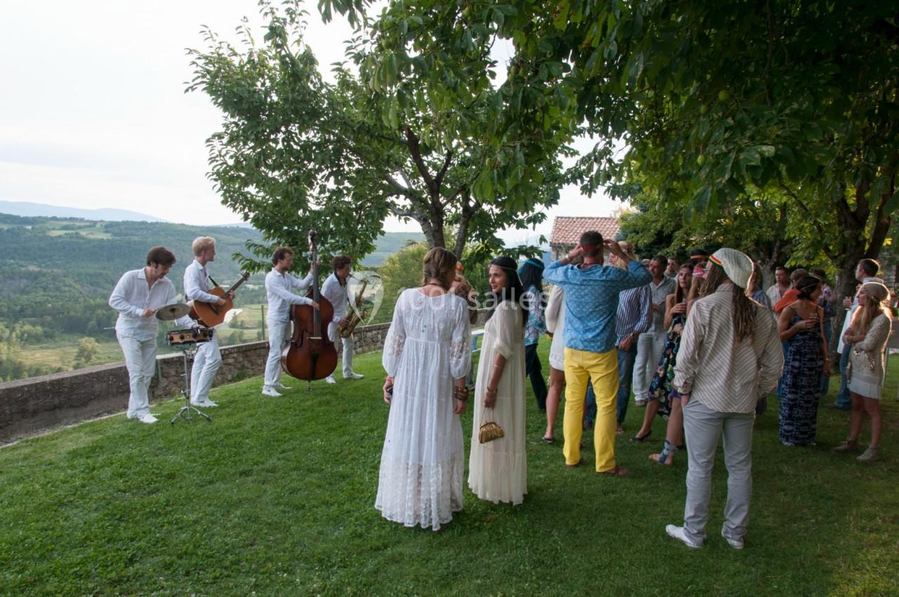 Un groupe de musiciens joue en plein air devant un public, dans un cadre verdoyant et ensoleillé.