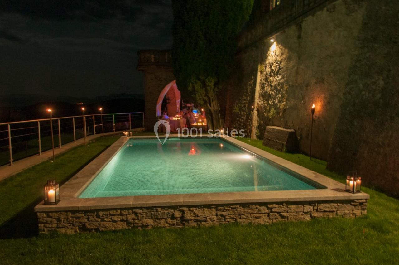 Piscine éclairée de nuit entourée de pelouse, avec des lanternes et un bâtiment en pierre en arrière-plan.
