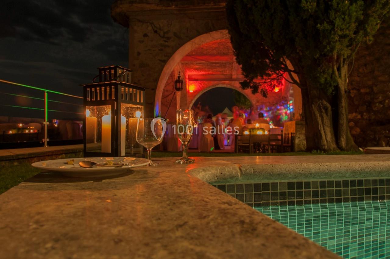 Table avec verres et bougies près d'une piscine, éclairée par des lumières rouges lors d'un événement nocturne en extérieur.