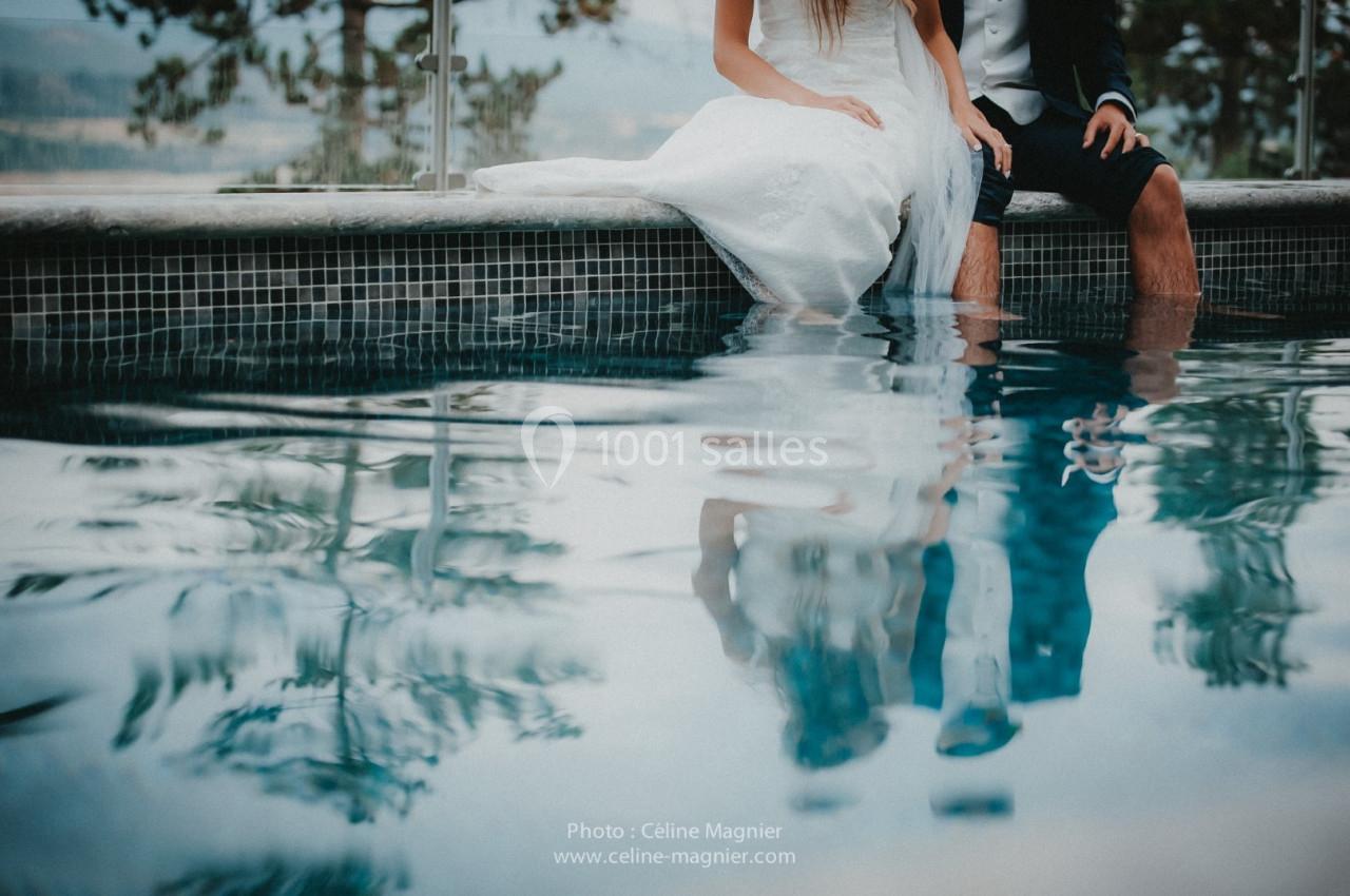 Un couple en tenue de mariage assis au bord d'une piscine, leurs reflets visibles dans l'eau calme.