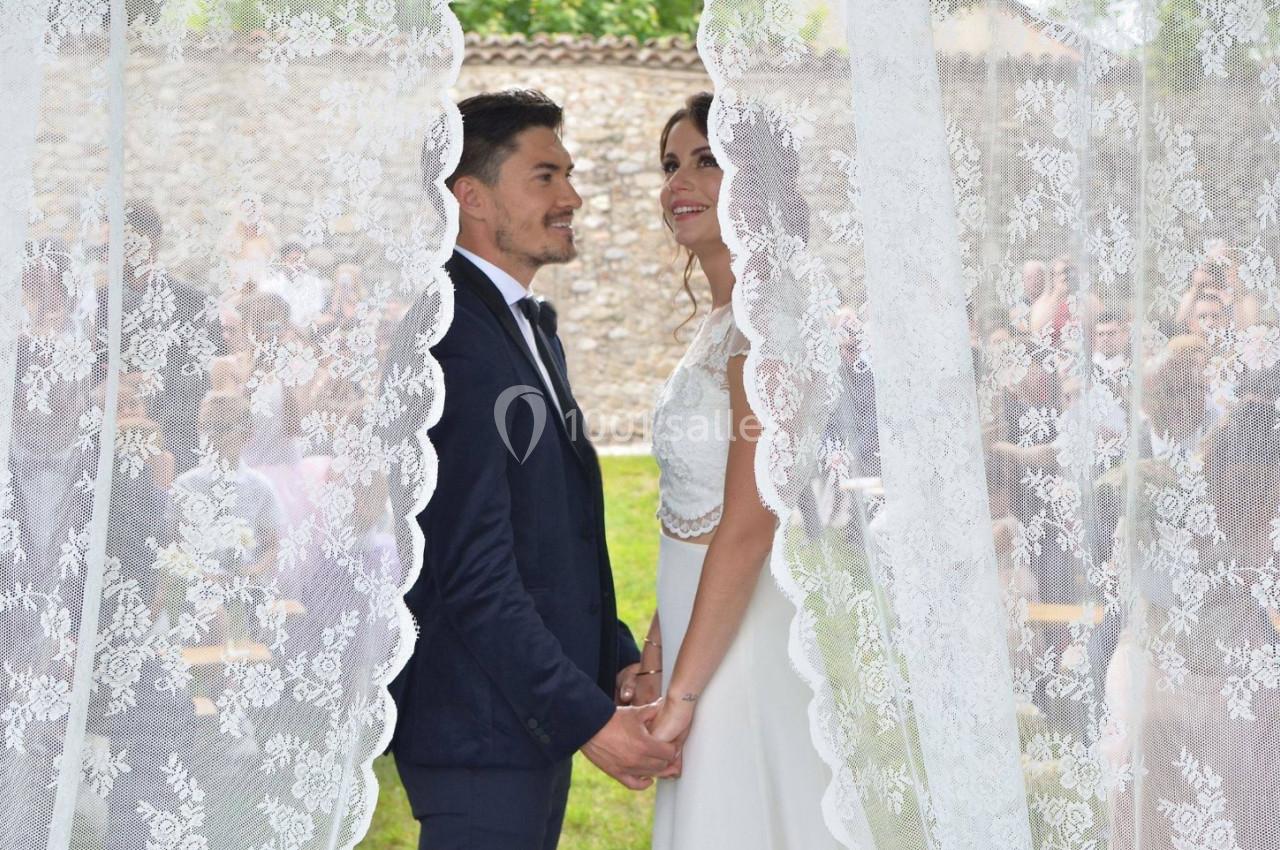 Un couple en tenue de mariage se tient la main, encadré par des rideaux en dentelle, avec des invités en arrière-plan flou.