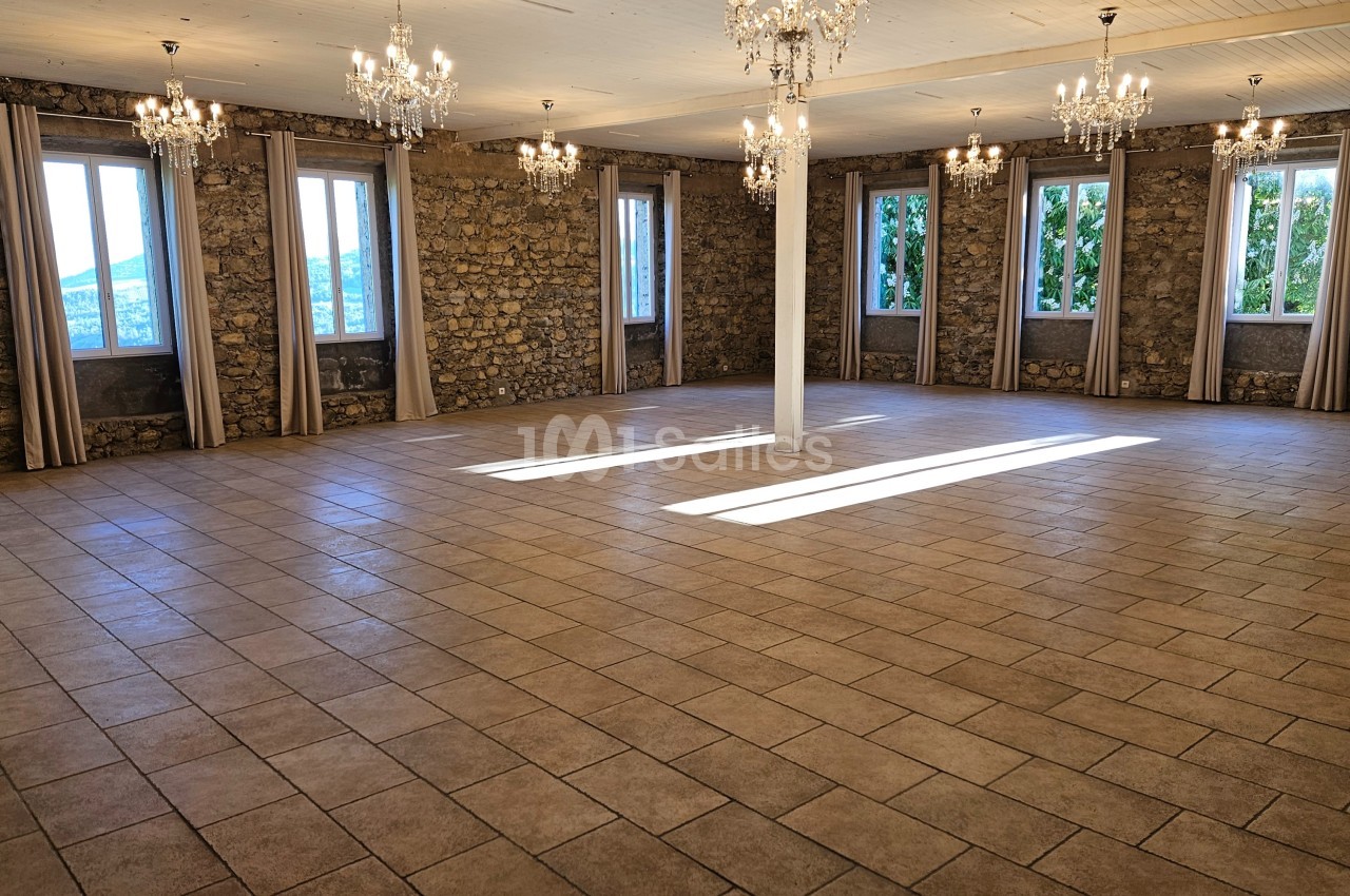 Salle spacieuse avec murs en pierre, carrelage au sol, grandes fenêtres et lustres suspendus.