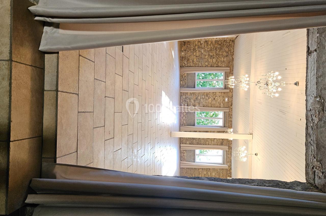 Salle lumineuse avec murs en pierre, carrelage beige, lustres suspendus et grandes fenêtres donnant sur l'extérieur.