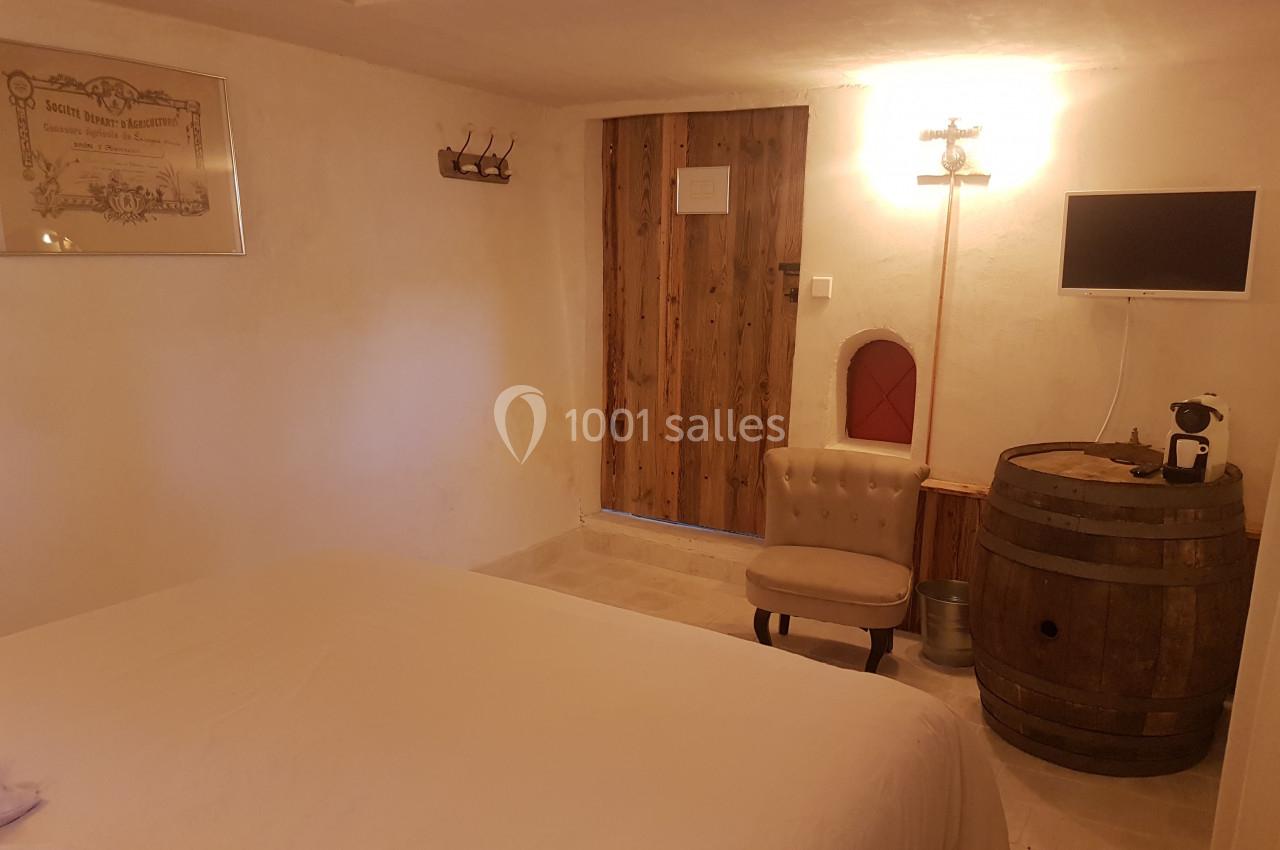 Chambre rustique avec lit double, mur en pierre, porte en bois, fauteuil, tonneau en guise de meuble et petite télévision.