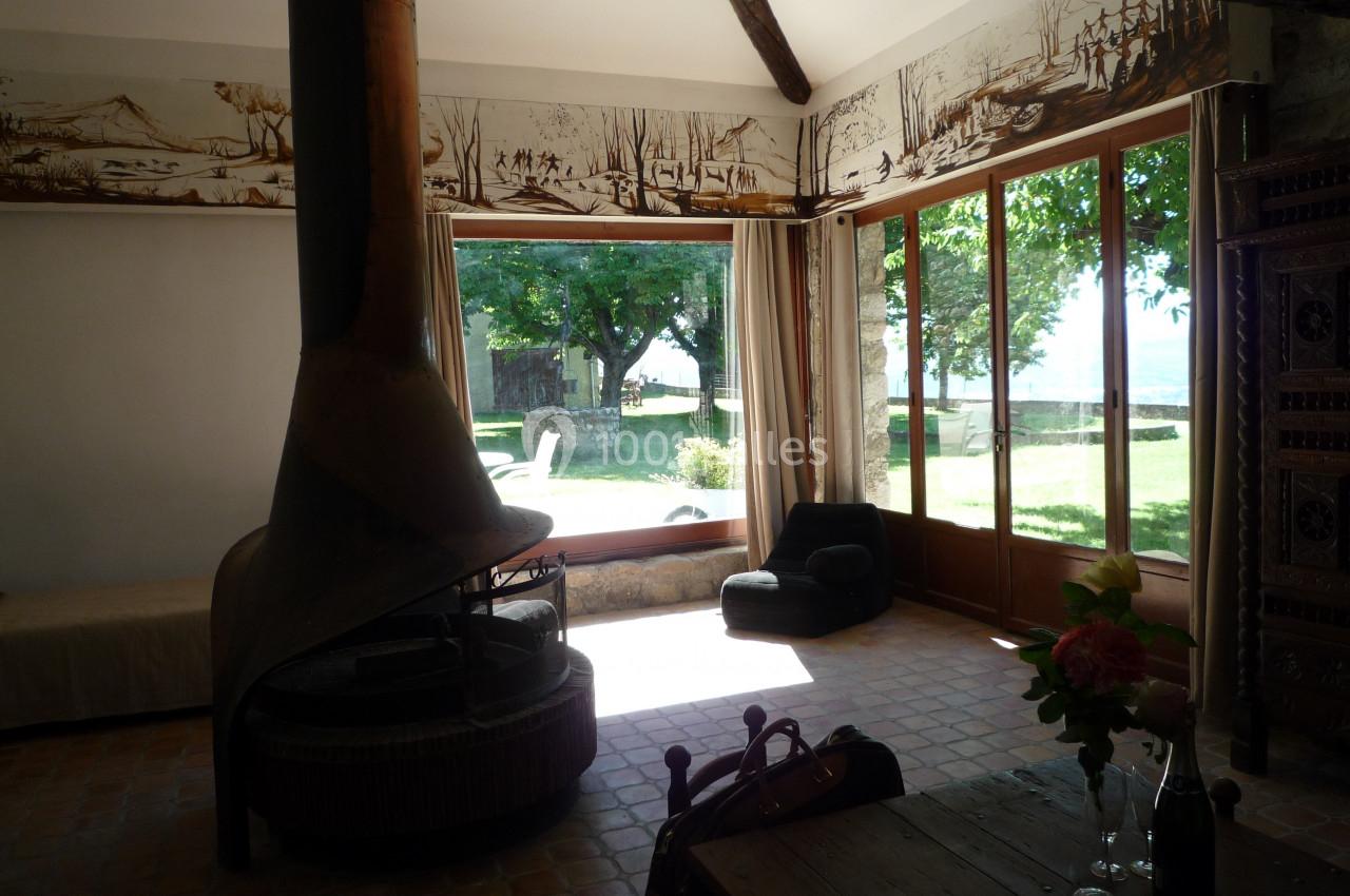 Salon lumineux avec cheminée centrale, grandes baies vitrées donnant sur un jardin arboré et mobilier rustique.
