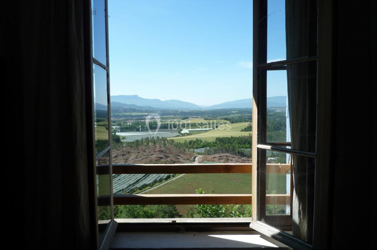 Vue depuis une fenêtre ouverte sur un paysage rural avec champs, collines et un ciel dégagé.
