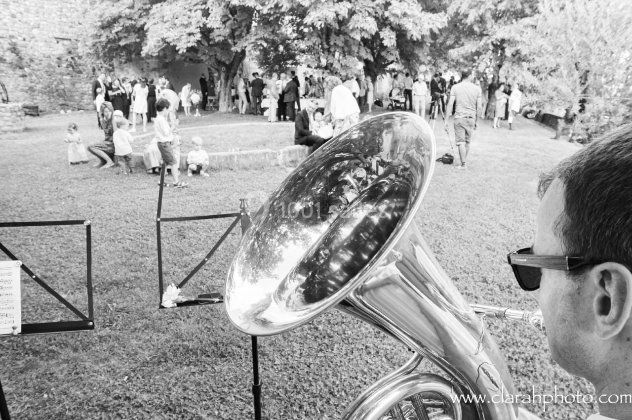 Un musicien joue du tuba lors d'un événement en plein air, avec des invités rassemblés sous des arbres en arrière-plan.