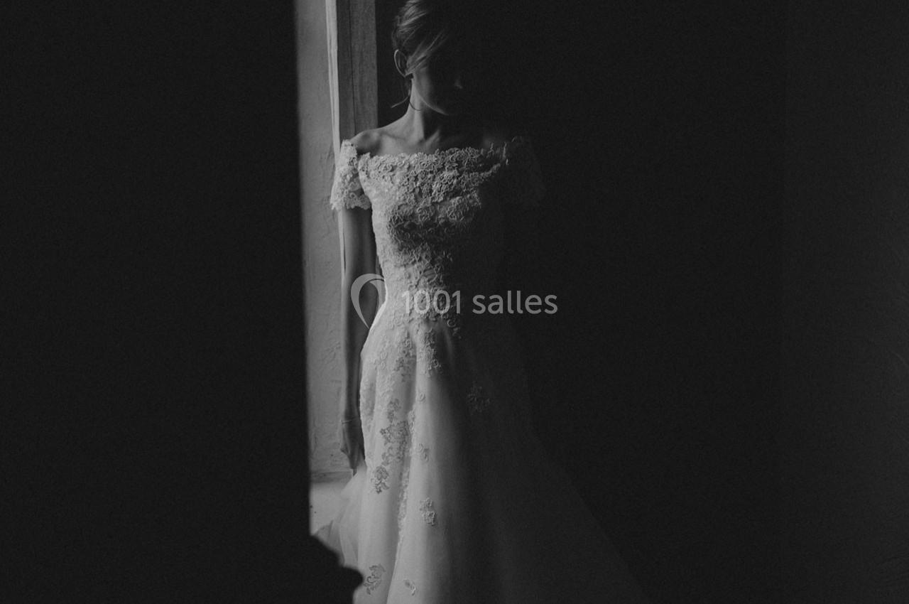 Femme en robe de mariée en dentelle, debout près d'une fenêtre dans une ambiance sombre et tamisée.