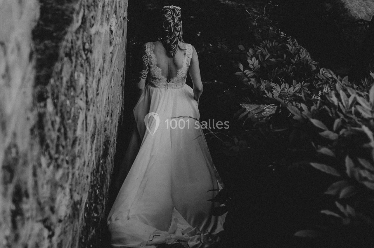 Une femme en robe de mariée vue de dos, marchant près d'un mur en pierre dans un environnement sombre et végétal.