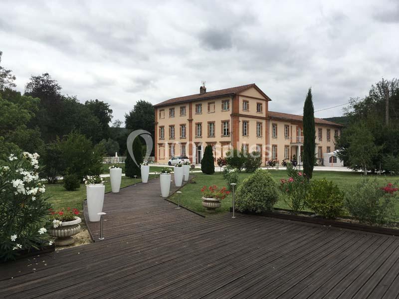 Allée en bois menant à un grand bâtiment ancien entouré de jardins fleuris sous un ciel nuageux.