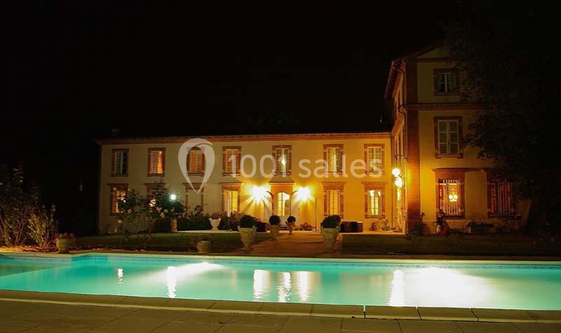 Façade éclairée d'une grande maison avec piscine illuminée au premier plan, dans un cadre nocturne.