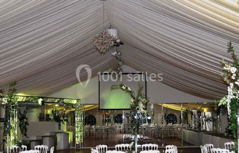 Intérieur d'une tente de réception décorée avec des lustres, des fleurs et des tables dressées pour un événement.