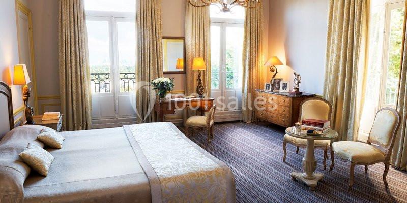 Chambre lumineuse avec lit double, bureau en bois, fauteuils et grandes fenêtres donnant sur un paysage verdoyant.