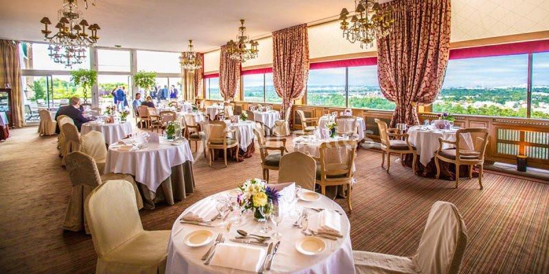 Salle lumineuse avec de grandes baies vitrées, tables dressées pour un repas, décorations élégantes et vue panoramique.