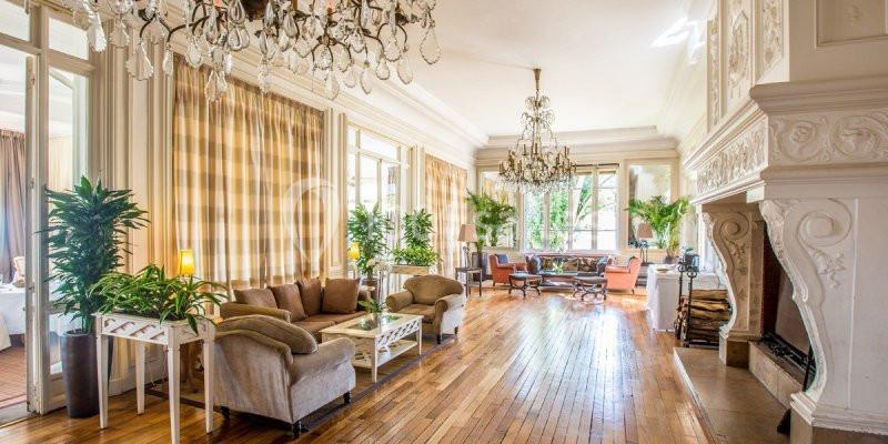 Salon lumineux avec parquet en bois, canapés, cheminées sculptées, lustres et grandes fenêtres donnant sur un jardin.