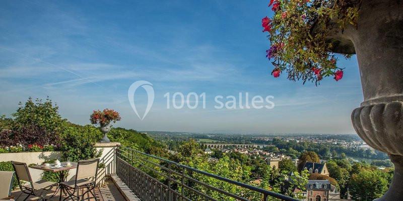Vue depuis une terrasse avec table et chaises, donnant sur un paysage verdoyant et une ville à l'horizon.