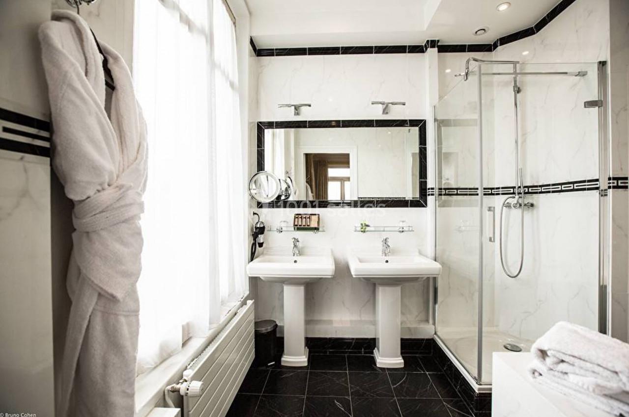 Salle de bain lumineuse avec deux lavabos, une douche vitrée, un peignoir suspendu et des murs en marbre blanc.