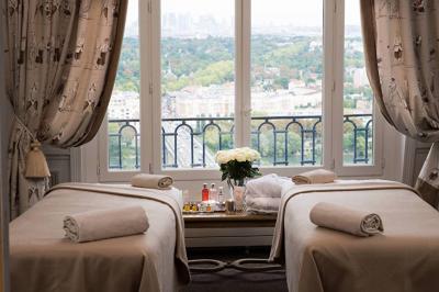 Deux tables de massage recouvertes de draps beiges, avec une vue sur un paysage urbain et verdoyant à travers une fenêtre.