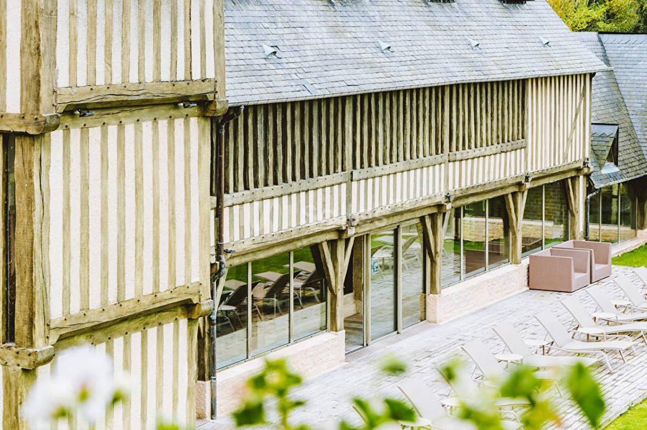 Façade en colombages d'un bâtiment avec terrasse en bois, chaises longues et baies vitrées donnant sur un espace vert.
