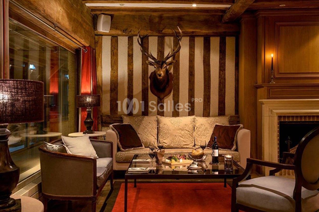 Salon chaleureux avec canapé, cheminée, tête de cerf au mur, table basse dressée et éclairage tamisé.
