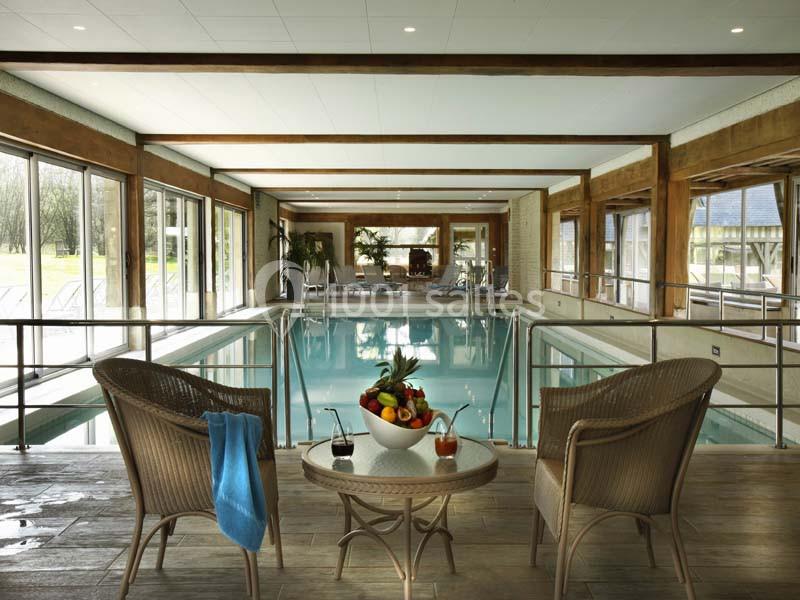 Piscine intérieure lumineuse avec chaises en osier, table garnie de fruits et grandes baies vitrées donnant sur l'extérieur.