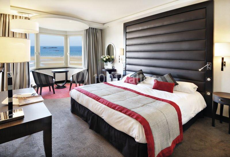 Chambre d'hôtel lumineuse avec un grand lit, coin salon, bureau et vue sur la mer par une large fenêtre.