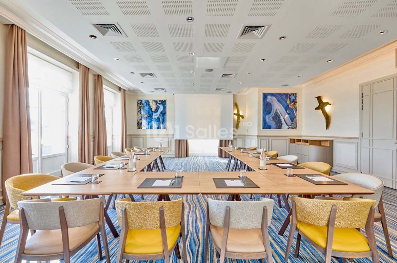Salle de réunion lumineuse avec tables disposées en U, chaises colorées, écran de projection et décorations murales.
