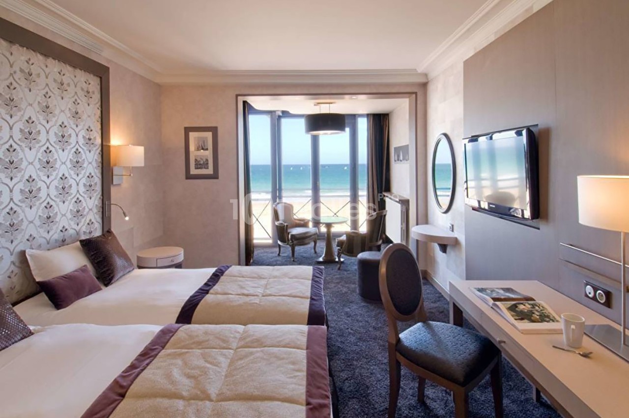 Chambre d'hôtel lumineuse avec deux lits, vue sur la mer, balcon, bureau et décoration élégante.