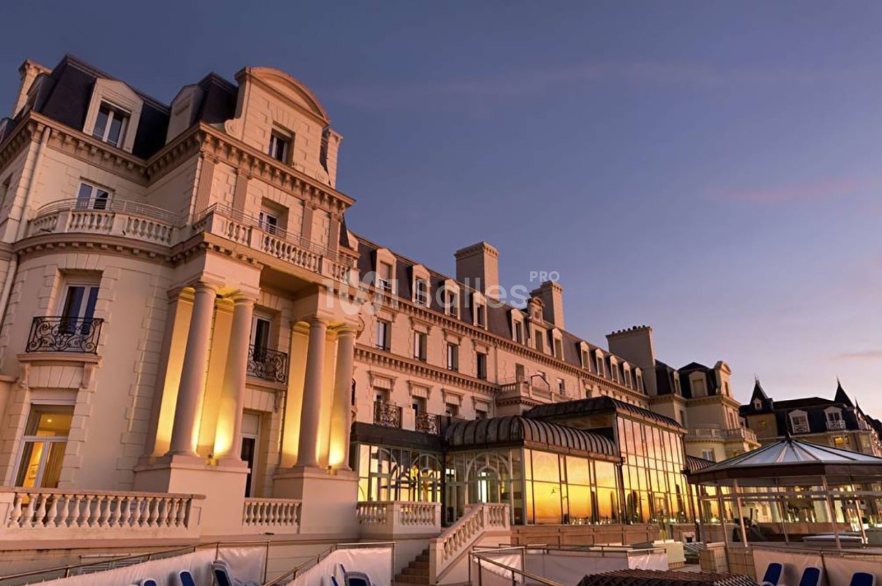 Façade d'un grand bâtiment historique éclairé au crépuscule, avec une verrière et une terrasse en premier plan.