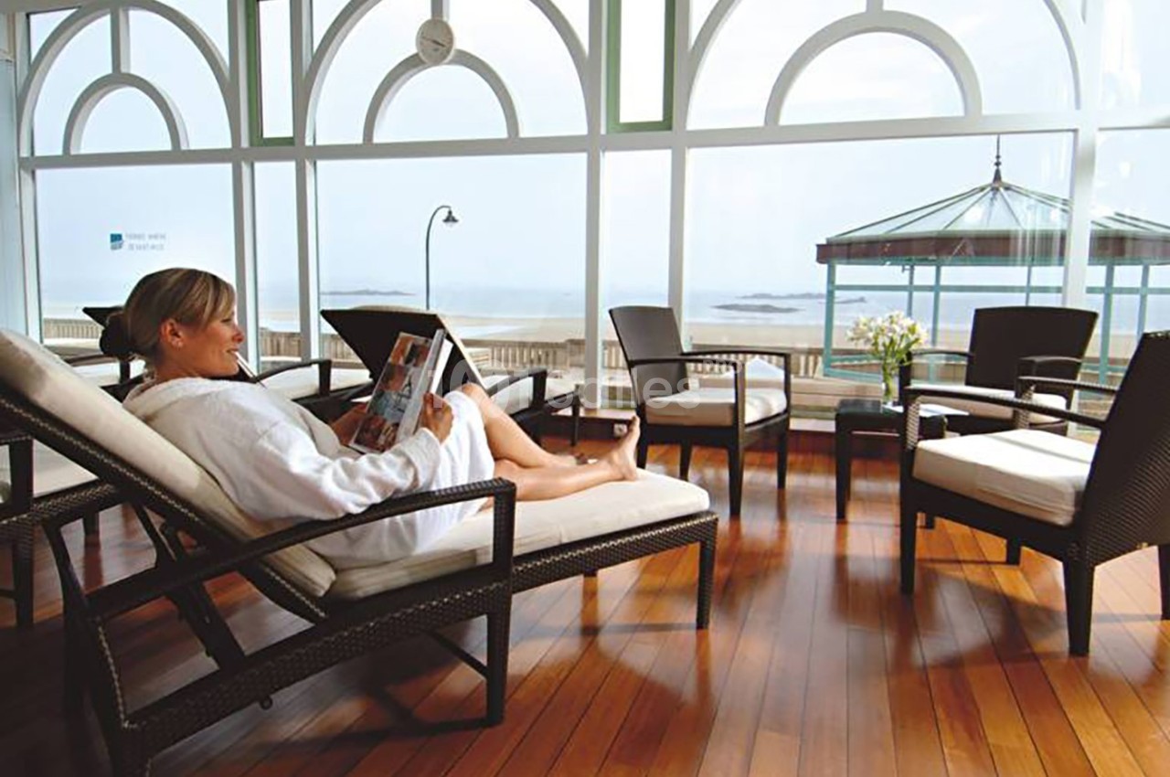 Femme en peignoir lisant un magazine sur une chaise longue dans une salle lumineuse avec vue sur la mer.