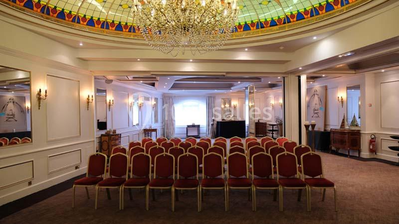 Salle de conférence élégante avec des rangées de chaises rouges, un lustre imposant et un plafond vitré coloré.